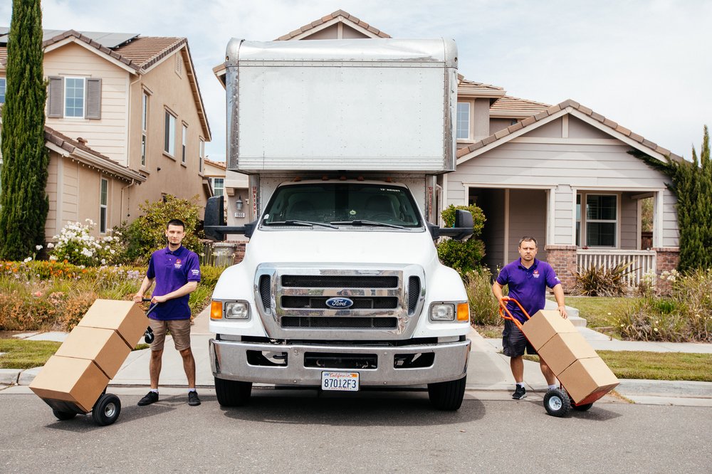 Two Double Move crew members with hand trucks of boxes standing on either side of a white Double Move box truck in a suburban driveway